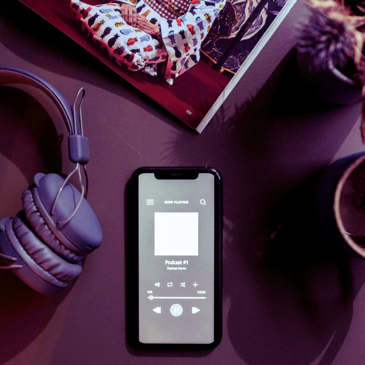 A flat lay image of a smartphone playing a podcast next to headphones and a mug.